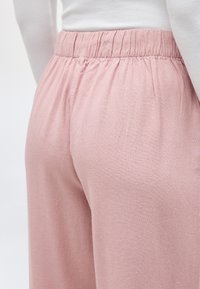 Κοντινό πλάνο ενός ατόμου που φοράει παντελόνι με ψηλή μέση και ελαστική ζώνη μέσης σε ανοιχτό ροζ χρώμα, συνδυασμένο με λευκό τοπ που είναι μπαλωμένο μέσα στο παντελόνι.