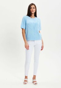 Light blue short-sleeve cardigan with buttons, made of soft material featuring a textured pattern, worn over white fitted pants and heeled sandals.