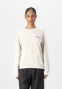 Sudadera beige en un material suave con puños y dobladillo acanalados. Presenta "STUDIO" bordado en rojo en la parte izquierda del pecho.