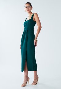 Turquoise nauwsluitende jurk met brede bandjes, vierkante halslijn en een voorsplit; gecombineerd met gouden sandalen met dunne bandjes en grote gouden oorbellen.