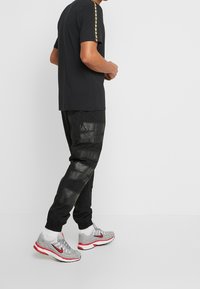 Man som bär svarta joggerbyxor med texturerade paneler, svart t-shirt med gul rand på ärmen, vita strumpor och grå sneakers med röda detaljer.