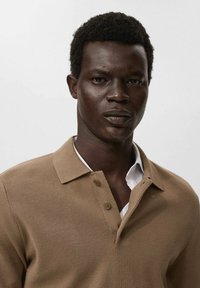 Man with short curly hair wearing a beige knit polo shirt over a white collared shirt, facing forward against a plain background.