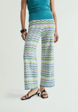 Mujer con pantalones de pierna ancha con patrón de zigzag en tonos pastel, top sin mangas verde azulado, pulsera negra y sandalias negras de tacón bajo, de pie sobre fondo blanco.
