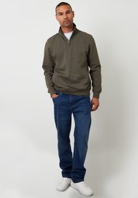Sudadera de cremallera color verde oliva con cuello alto, combinada con jeans de mezclilla azules y zapatillas blancas. Textura suave y ajuste relajado.