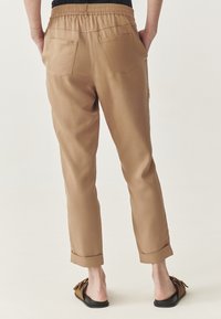 Tan tapschoenen met een elastische tailleband, twee achterzakken en opgerolde manchetten bij de enkels, gemaakt van soepel, lichtgewicht materiaal.