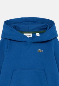 Sweatshirt azul com um bolso canguru e punhos canelados. O interior tem um forro verde, e um logo de crocodilo bordado está posicionado no peito.