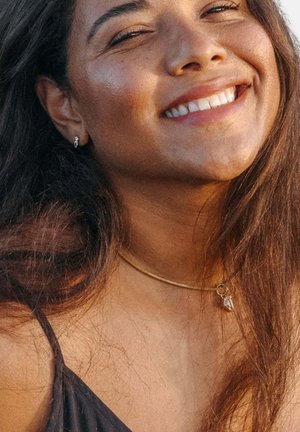 Junge Frau mit langen dunklen Haaren, die lächelt, trägt kleine silberne Ohrringe, eine dünne goldene Halskette mit einem Kristallanhänger und ein schwarzes Top mit dünnen Trägern.