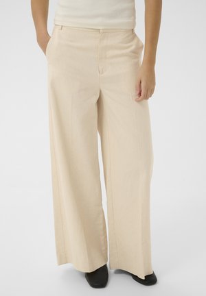 Kobieta nosząca beżowe spodnie z wysokim stanem i szerokimi nogawkami z bocznymi kieszeniami oraz czarne buty z kwadratowymi noskami, stojąca na jednolitym tle.