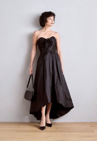 Juoda be petnešų suknelė su gėlių akcentu, klostuota sijonų, aukščiau-žemiau hemline, derinama su juodais smailiais aukštakulniais ir mažomis juodomis rankinėmis.