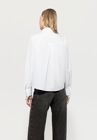 White long-sleeve shirt with back pleat detail, buttoned cuffs, and rounded hem, paired with dark, wide-leg jeans.