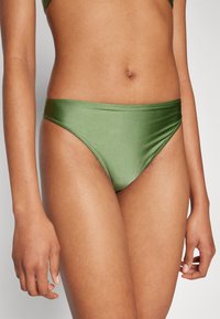 Costumi da bagno verde in basso realizzati in materiale lucido, caratterizzati da un design a vita bassa e copertura minima. Texture liscia con cuciture pulite.