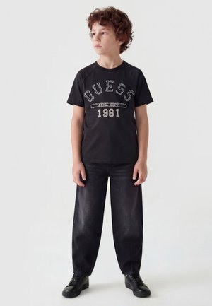 Chłopiec z kręconymi włosami, ubrany w czarną koszulkę "GUESS 1981", czarne spodnie i czarne trampki, stoi i patrzy na bok.