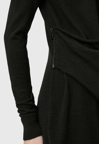 Gros plan sur un vêtement noir à manches longues avec une poche zippée diagonale sur le côté, montrant un tissu texturé et des poignets ajustés.