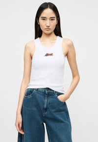 Νεαρή γυναίκα με μακριά σκουρόχρωμα μαλλιά φοράει λευκό αμάνικο τοπ με μικρό σχέδιο ενός τρέχοντα αλεπού και φαρδιά μπλε τζιν, με το ένα χέρι στην τσέπη.