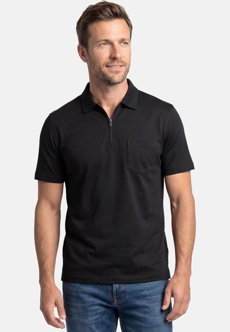 Mann trägt ein schwarzes kurzärmeliges Polohemd mit einem vorderen Reißverschluss und Brusttasche, kombiniert mit blauen Jeans, vor einfachem Hintergrund.