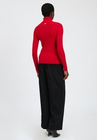 Maglione a collo alto rosso con texture a costine, abbinato a pantaloni neri larghi e tacchi alti neri. Design minimalista, senza pattern visibili.