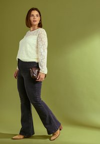 Top de encaje blanco de manga larga con textura ondulada, combinado con jeans oscuros de campana y botas de tacón de tobillo en color beige. Sosteniendo un pequeño bolso de mano marrón.