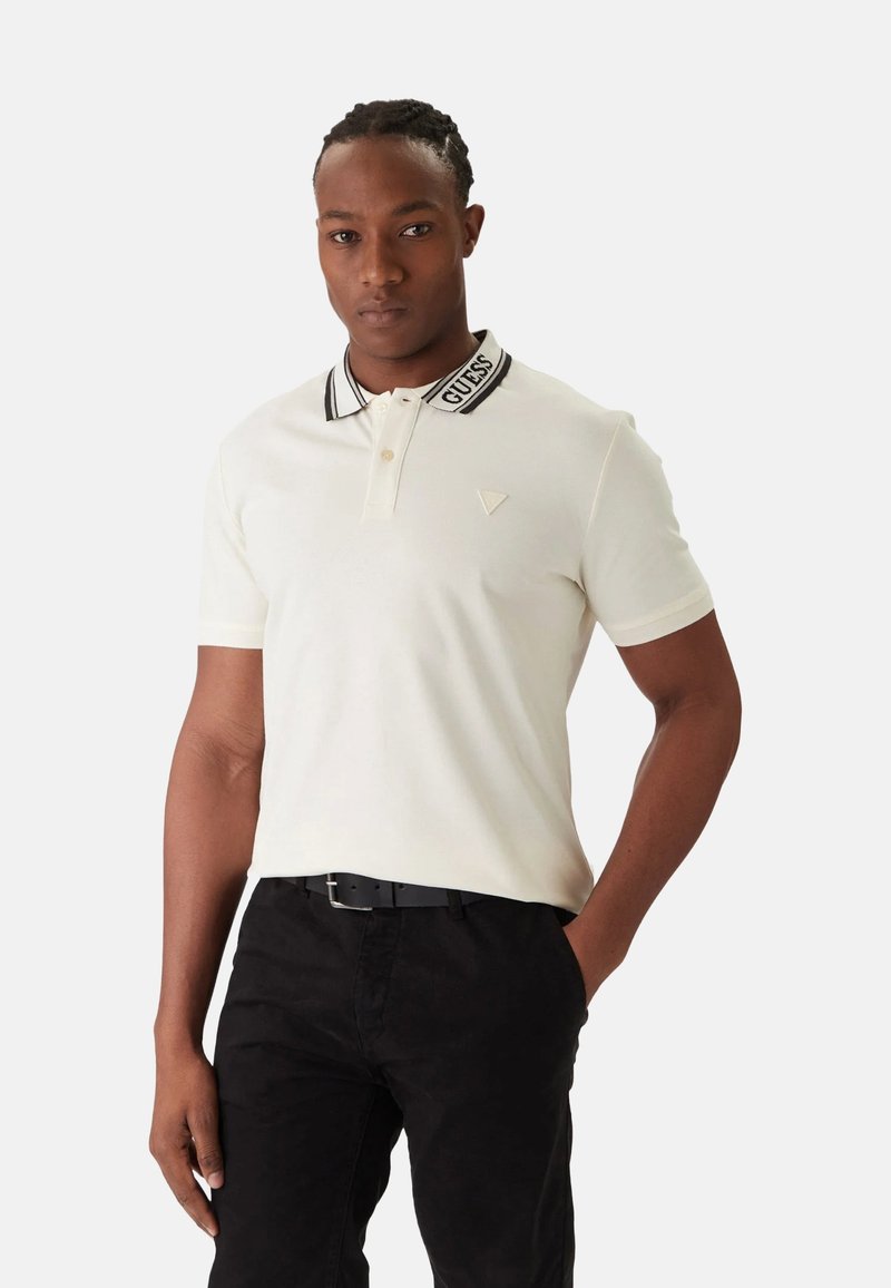 Uomo che indossa una polo bianca con la scritta "GUESS" sul colletto e pantaloni neri, in piedi con una mano in tasca davanti a uno sfondo semplice.