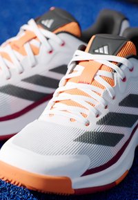 Witte sportschoenen met oranje en grijze accenten, voorzien van een gestructureerde mesh bovenkant, witte veters en een opvallend gestreept ontwerp op de zijkanten.