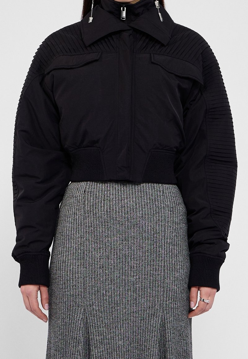Femme portant une veste matelassée noire avec des manches côtelées et un col montant sur une jupe en tricot côtelé grise.