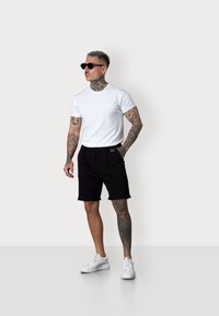 White short-sleeve t-shirt, black shorts, and white sneakers. Model has tattoos, wearing black sunglasses. Clean lines and minimal design.