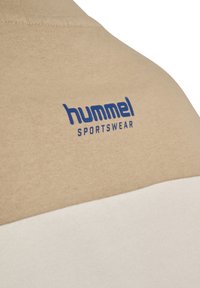 Beige bomulds sportswear med en hvid nedre del. Har et blåt Hummel-logo og tekst på den øvre del, der viser en glat tekstur og syninger.