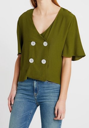 Kvinde iført olivengrøn kortærmet bluse med fire hvide knapper, kombineret med blå denimjeans, står med afslappede arme.