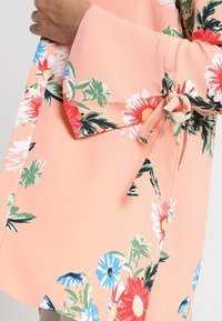 Blommig blus i mjuk persikofärgad tyg med luftiga ärmar, prydd med röda, vita och blå blommor. Inkluderar ett midjeband för justerbar passform.