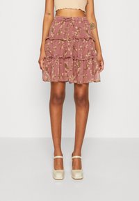 Femme portant une jupe à volants rose à fleurs avec une taille à cordon et des chaussures Mary Jane de couleur crème, adossée à un fond uni.