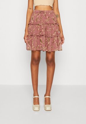 Femme portant une jupe à volants rose à fleurs avec une taille à cordon et des chaussures Mary Jane de couleur crème, adossée à un fond uni.
