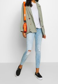 Blazer a cuadros verdes con botones marrones, camiseta blanca, jeans skinny rasgados en azul claro y zapatos negros sin cordones. Bolso cruzado naranja.