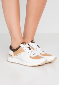 Bílé a krémové sportovní tenisky s hnědým leopardím vzorem, šněrovací design, perforované panely a zlaté detaily na patě.