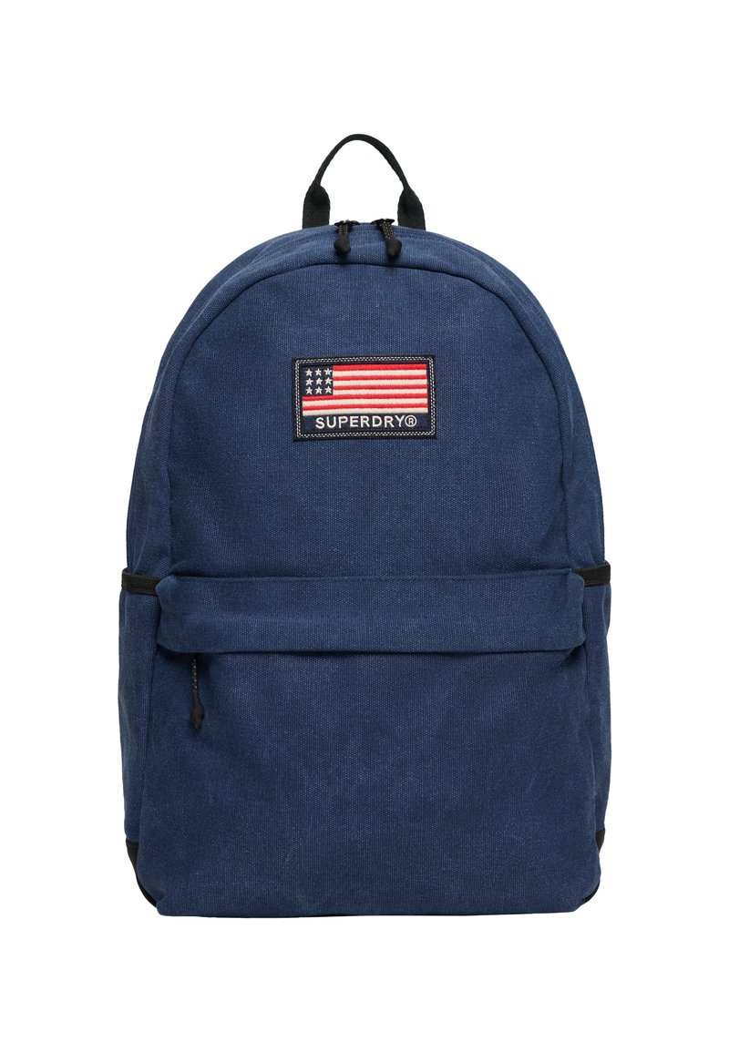 Granatowy plecak wykonany z tkaniny, z przednią kieszenią, czarnymi okuciami oraz prostokątną naszywką z amerykańską flagą i logo "SUPERDRY".