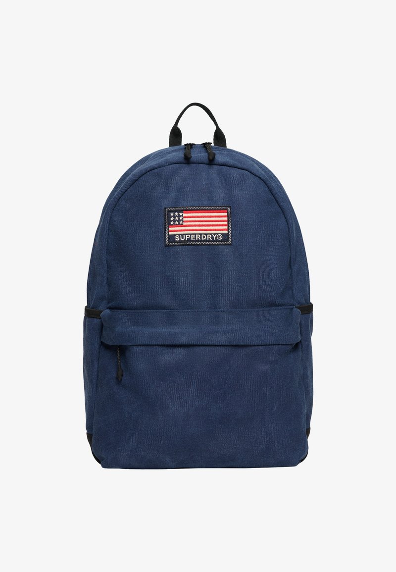Granatowy plecak wykonany z tkaniny, z przednią kieszenią, czarnymi okuciami oraz prostokątną naszywką z amerykańską flagą i logo "SUPERDRY".