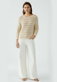 Beige gestrickter Pullover mit offener Maschenstruktur und horizontalen Streifen, kombiniert mit weißen weit geschnittenen Hosen und braunen Sandalen. Dezente Ohrringe ergänzen das Outfit.