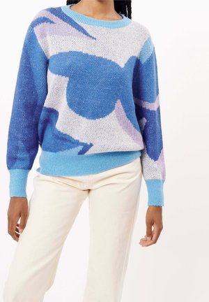 Osoba nosící svetřík s abstraktním vzorem v pastelových barvách světle modré, fialové a bílé s světle modrými manžetami a krémové kalhoty, stojící před jednobarevným pozadím.