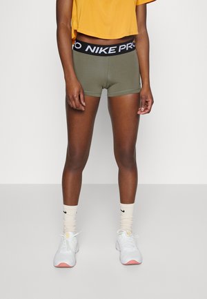 Person trägt olivgrüne Nike Pro Sportshorts, ein gelbes kurzes Top, cremefarbene Nike-Socken und weiße Turnschuhe und steht vor einem einfarbigen Hintergrund.