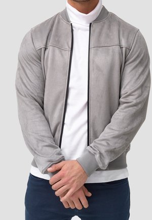 Mężczyzna w jasnoszarej zamszowej kurtce bomber na białym golfie z splecionymi rękami, na jednolitym jasnoszarym tle.