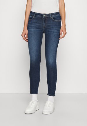 Persoon draagt donkerblauwe strakke spijkerbroek, witte sokken, witte sneakers en een wit kort mouwtopje dat in de broek is gestopt.