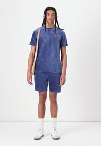 T-shirt en velours côtelé bleu et short assorti avec un petit logo. Le modèle porte un sac bandoulière beige et des chaussettes blanches à la cheville avec des baskets.