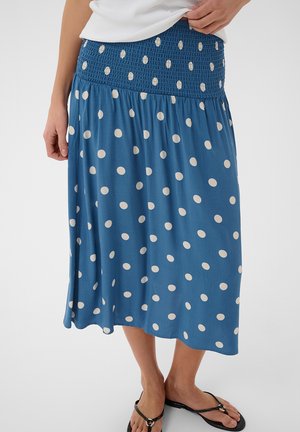 Blauer Midirock mit weißen Punkten, Raffung am Bund, getragen mit weißem Oberteil und schwarzen Zehenstegsandalen an einer stehenden Person.