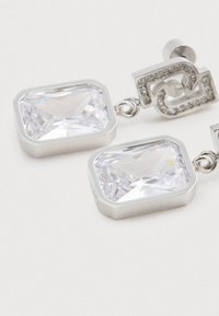 Sudraba krāsas auskari ar lieliem taisnstūra skaidriem kristāliem un mazākiem kristāliem, kas rotā augšējo ģeometrisko dizainu.