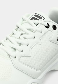 Közelkép egy fehér tornacipőről, amelynek texturált szövete, sima burkolatai, fehér fűzői és a nyelvén egy fekete logó része látható.