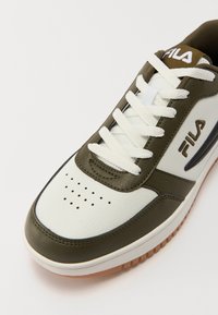 White and olive green sneaker with perforated toe, textured leather, flat white laces, and rubber outsole. Features Fila branding on the side.