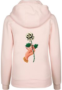 Rózsaszín kapucnis pulóver, hátulján egy arany virágot tartó kéz grafikájával, zöld levelekkel. Puha anyag, laza szabású.