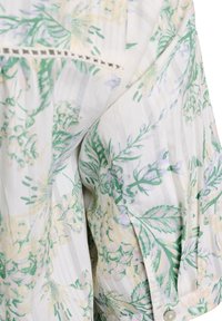 Blouse florale en tissu doux, mettant en avant des motifs verts et jaunes sur un fond rayé blanc. Comprend une bordure décorative au niveau de l'encolure.