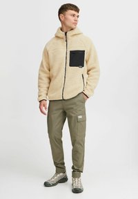 Beige fleecejakke med en svart lommepakke, glidelås foran og foret hette; kombinert med olivenfargede cargo-bukser og grå vandresko.