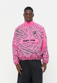 Nike Air lightweight zip-up jacket featuring a pink and gray geometric pattern, elastic cuffs, and a logo patch on the chest.