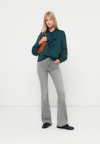 Blusa turchese con scollatura arricciata e maniche trasparenti, abbinata a jeans svasati grigio chiaro e scarpe nere. Una borsa a tracolla marrone è tenuta in mano.