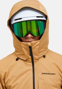 Giacca impermeabile beige con collo alto, cerniere nere e logo "PeakPerformance". Indossata con casco bianco e occhiali con lenti verdi.